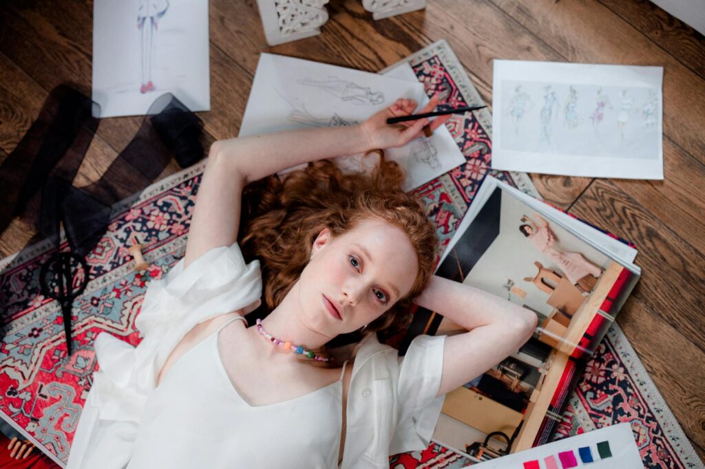 A redheaded woman relaxes indoors with her fashion sketches and designs.