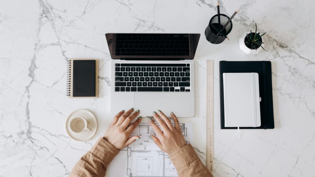 Top view of a stylish home office desk with a laptop, planner, and coffee cup, showing hands on a blueprint. - Tecno Pova 7 Neo 4G Design & Features Leaked Online